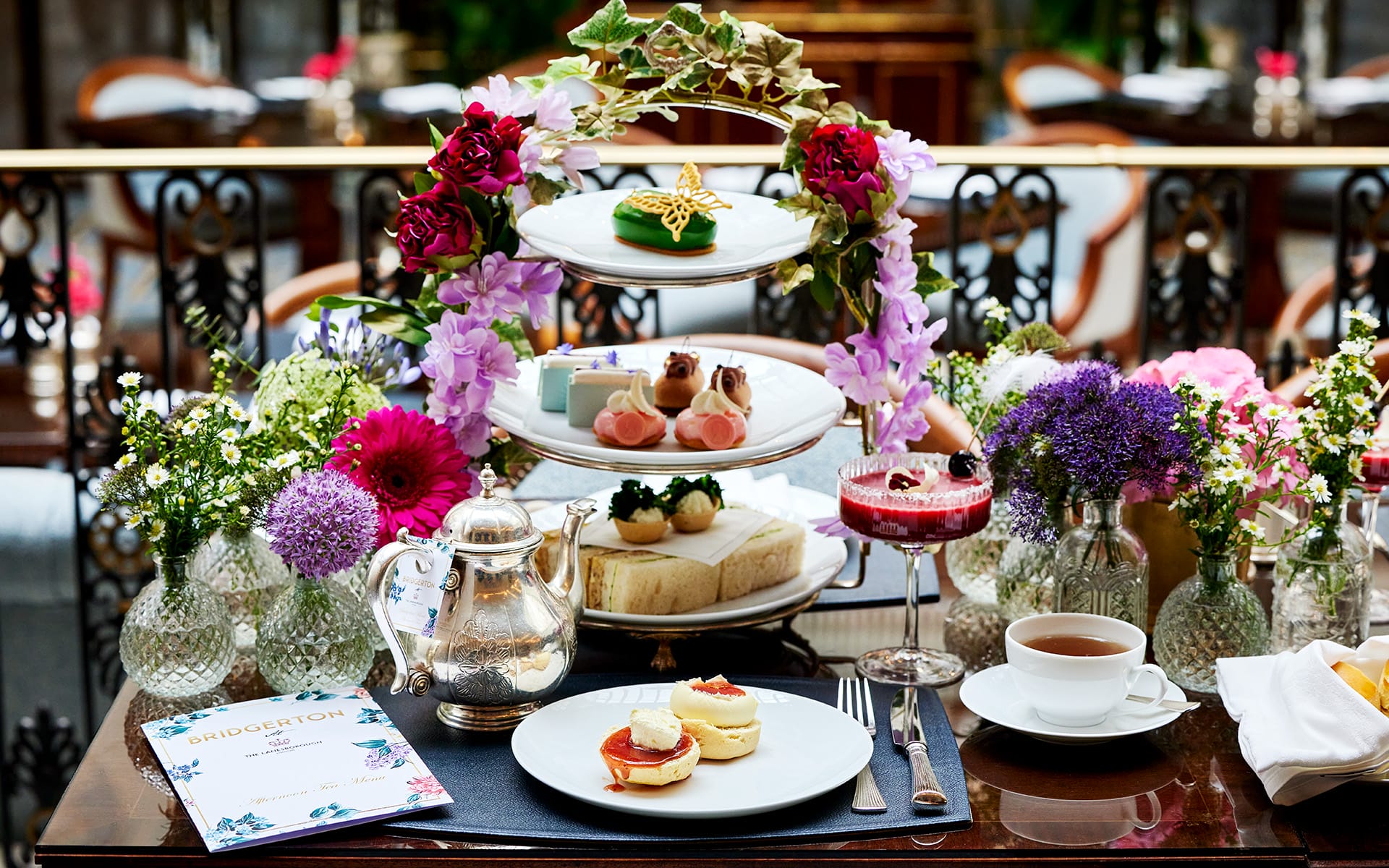 Afternoon tea setup with tiered tray of pastries, floral arrangements, teapot, and menu on a decorated table.