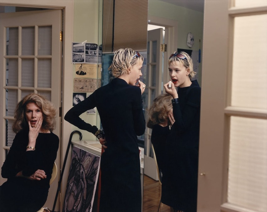 Woman applying lipstick in front of a mirror while another woman sits nearby, both in a room with various wall decorations.