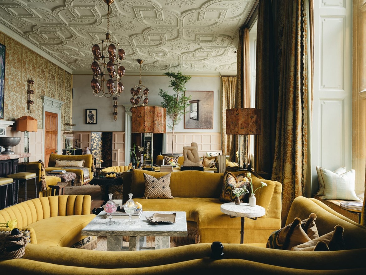 Elegant living room with vintage decor, ornate ceiling, mustard yellow sofas, and large windows letting in natural light.