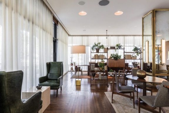 Modern living room with wooden floor, green armchairs, large windows, and plants on shelves, creating a cozy atmosphere.