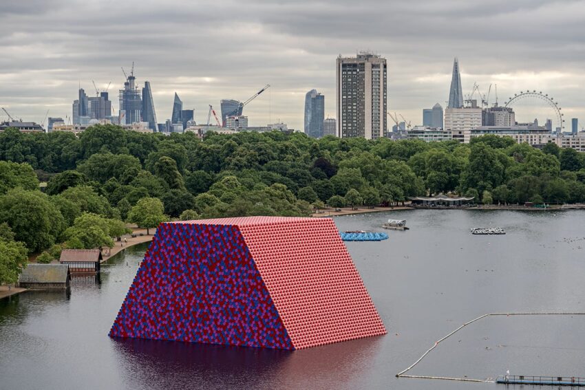 Colorful floating art installation on a lake with a city skyline and greenery in the background.