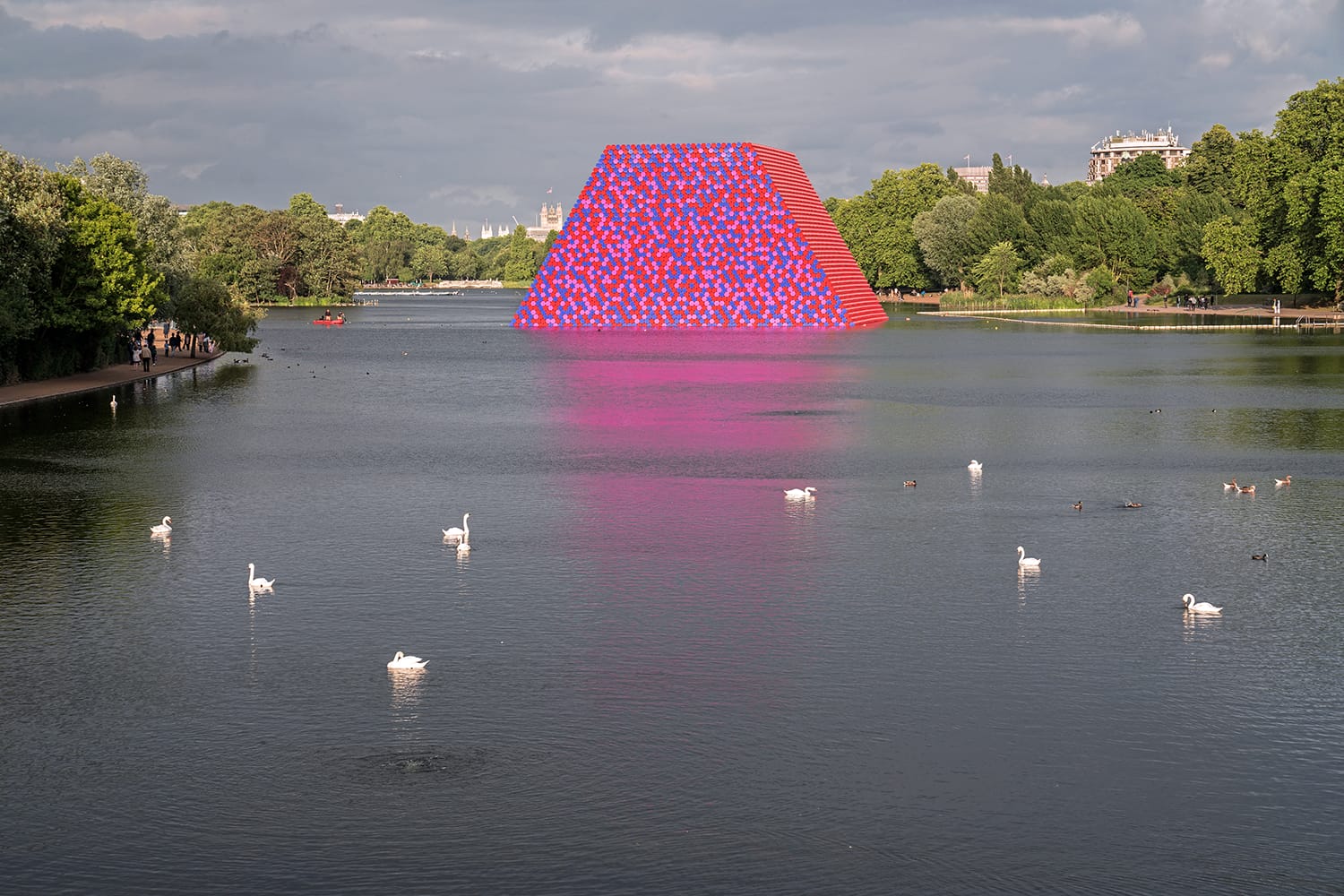 Colorful floating pyramid structure on a lake with swans in the foreground and trees in the background.