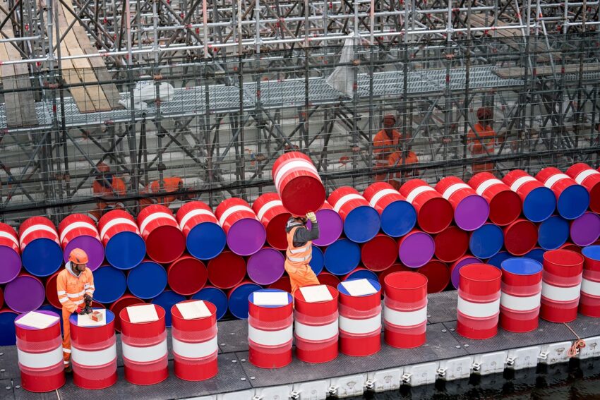 Workers stacking colorful barrels on a metal scaffold beside water, with multiple red, blue, and pink barrels in the background.