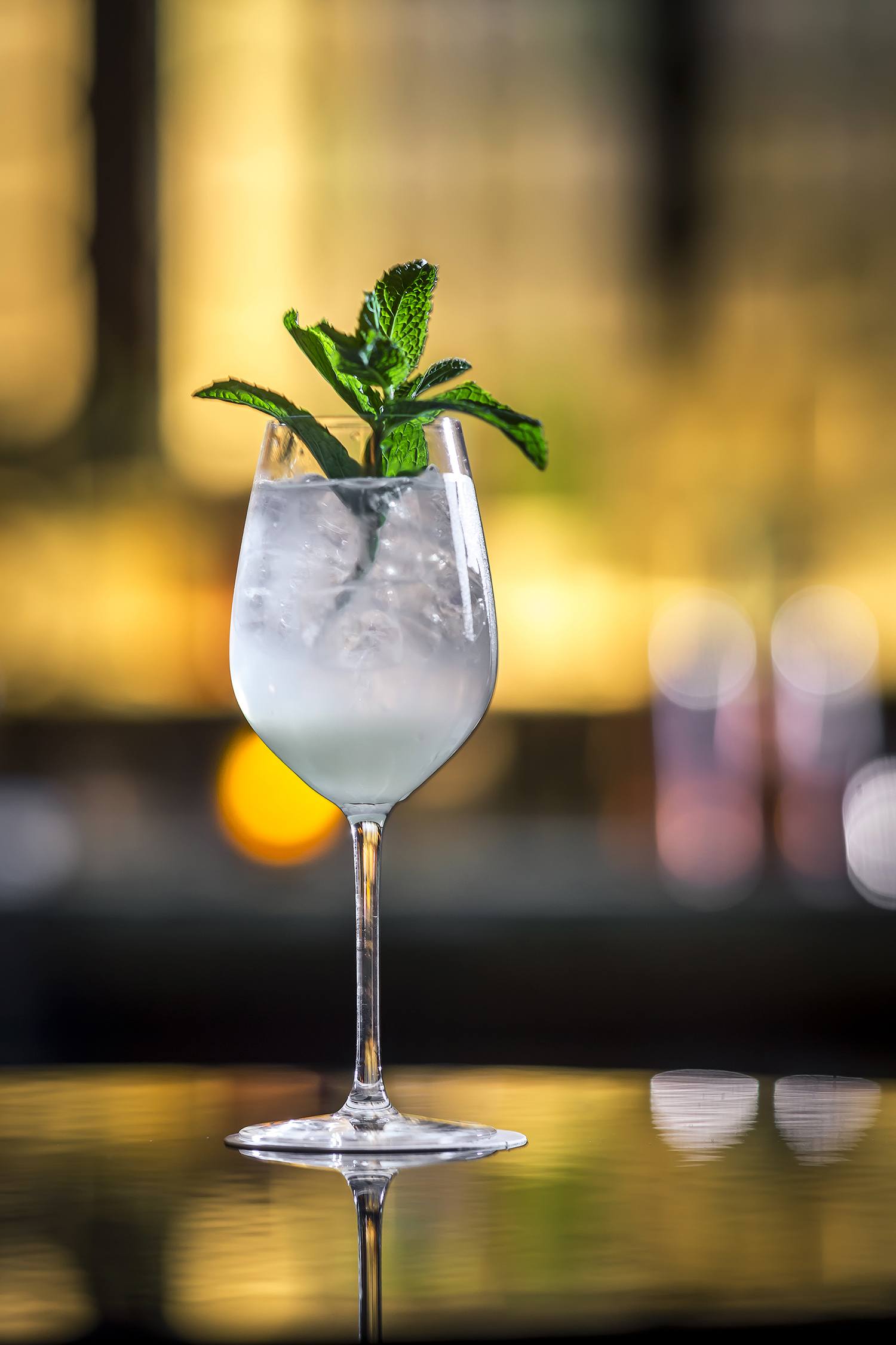 A chilled cocktail in a wine glass garnished with fresh mint leaves, set against a blurred background.