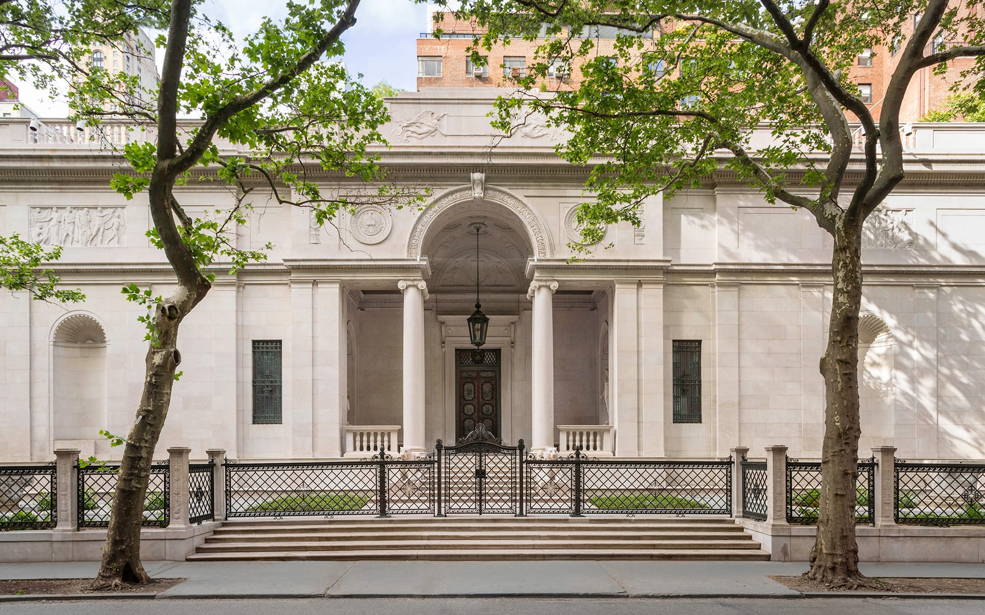 Historic white stone building with arched entrance, ornate carvings, and trees framing the entrance, seen from the street sidewalk.