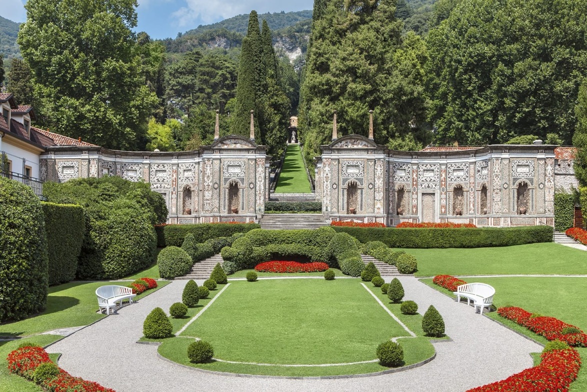 Italian villa garden with ornate stone wall, lush greenery, vibrant red flowers, and symmetrical pathways on a sunny day.