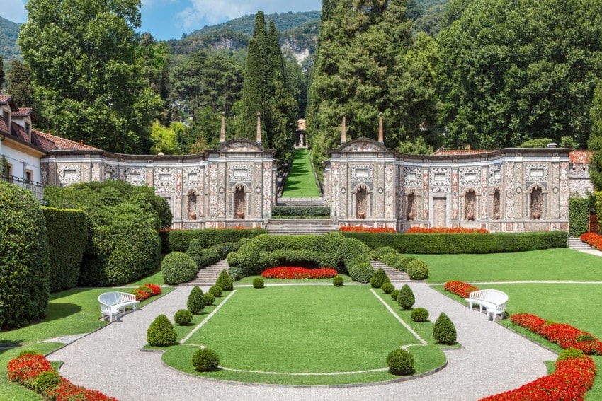 Elegant Italian garden with manicured lawns, vibrant red flowers, stone architecture, and lush green trees in the background.