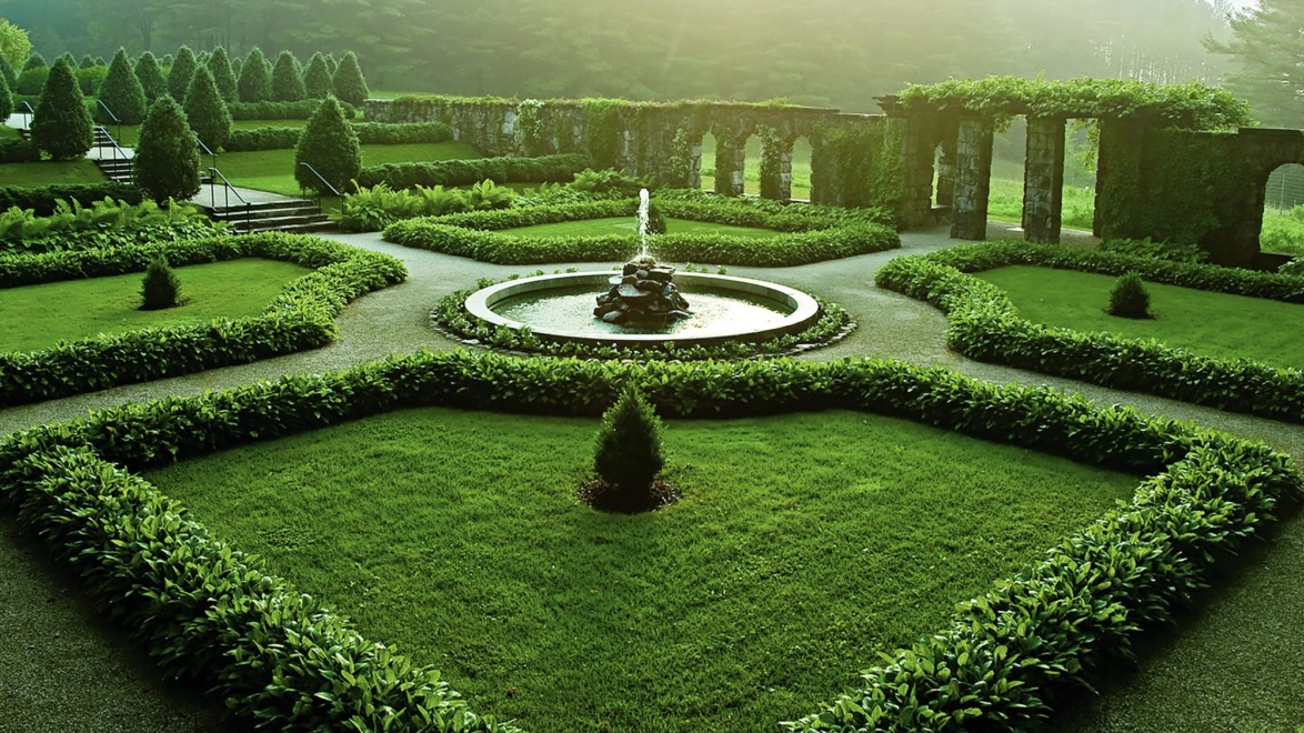 Formal garden with a central fountain surrounded by manicured hedges and lush green lawn, illuminated by soft sunlight.