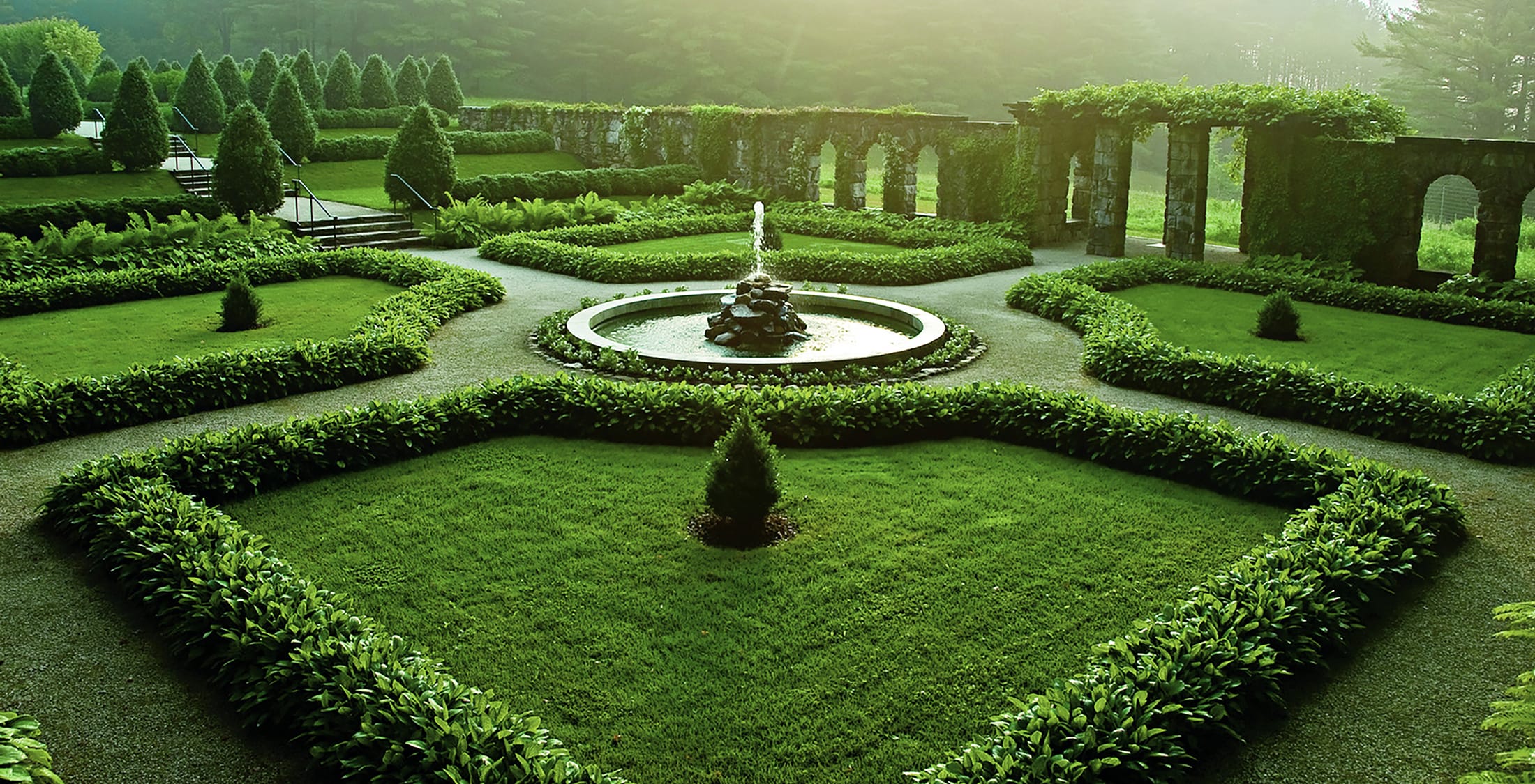 Formal garden with a central fountain surrounded by manicured hedges and lush green lawn, illuminated by soft sunlight.