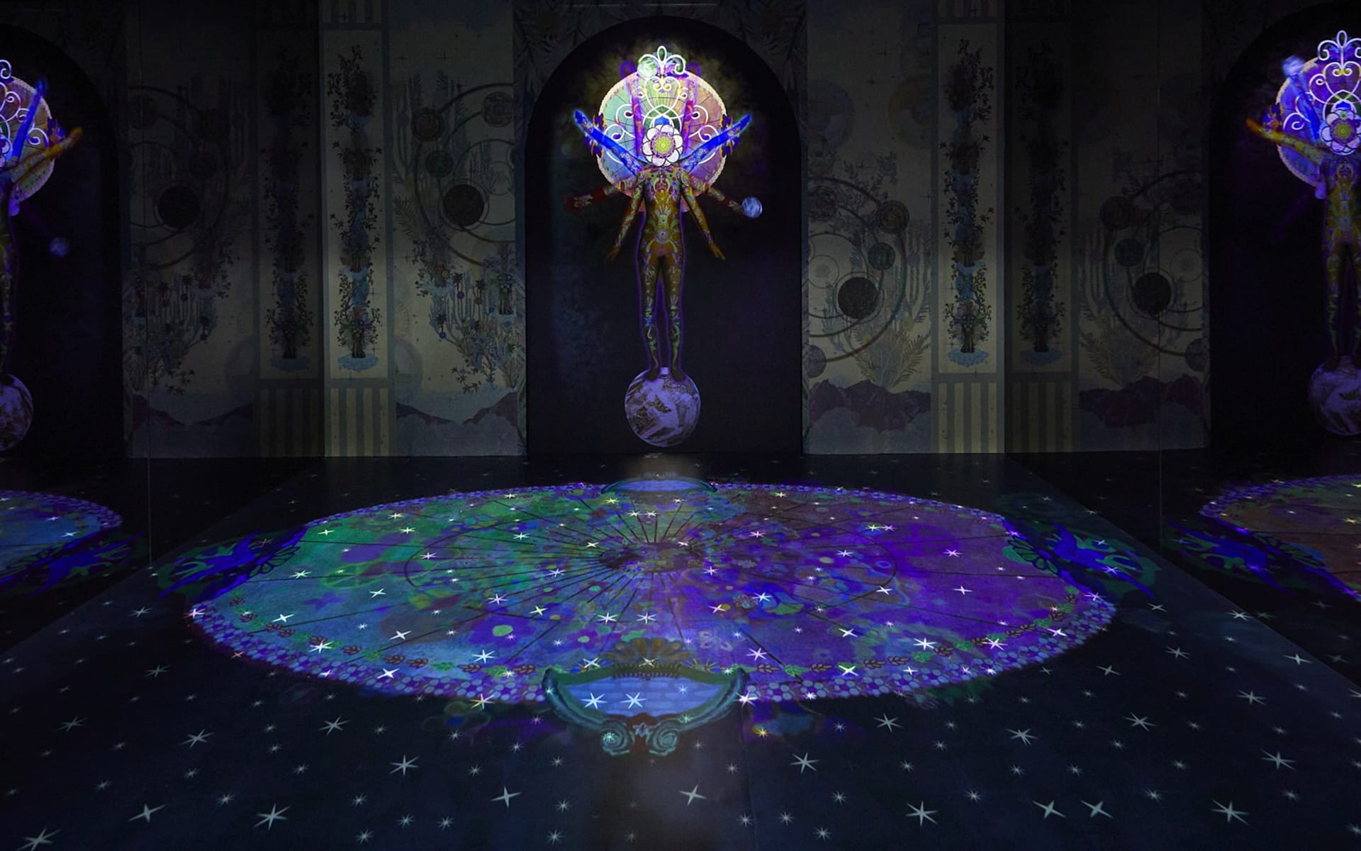 Colorful digital art projection on a dark surface featuring intricate patterns and an illuminated figure above a globe.