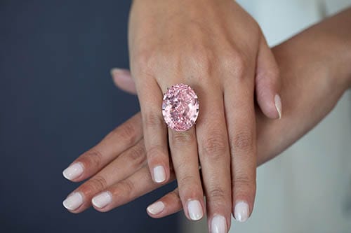 A hand displaying a large, oval pink diamond ring against a neutral background.