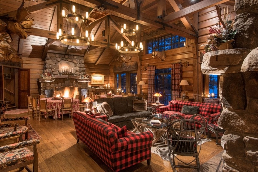 Cozy rustic cabin living room with plaid sofas, stone fireplace, wooden beams, antler chandeliers, and deer mounts.