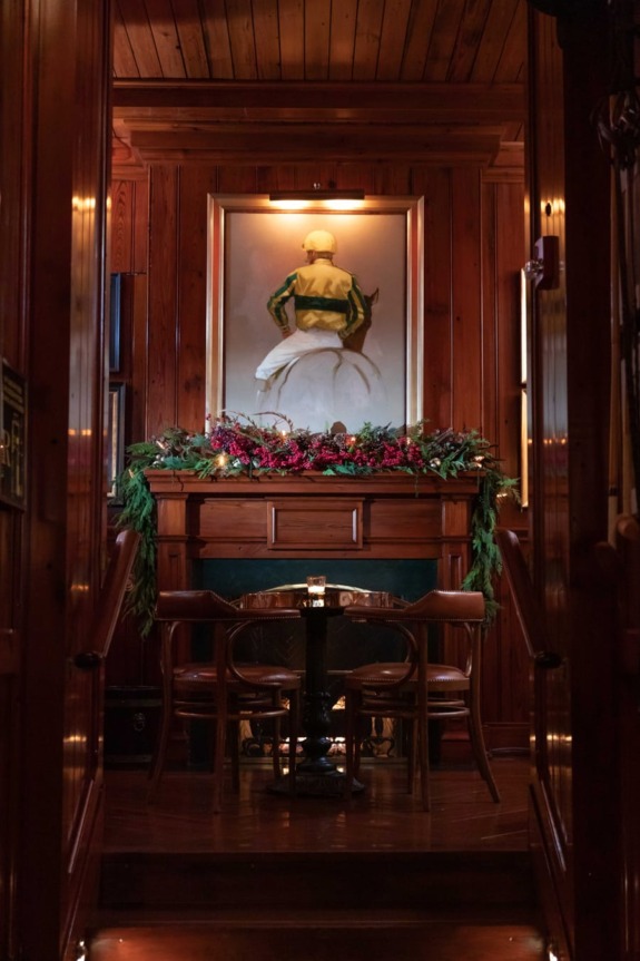 Cozy wooden dining area with a painting of a jockey, surrounded by festive garland above a small table and chairs.