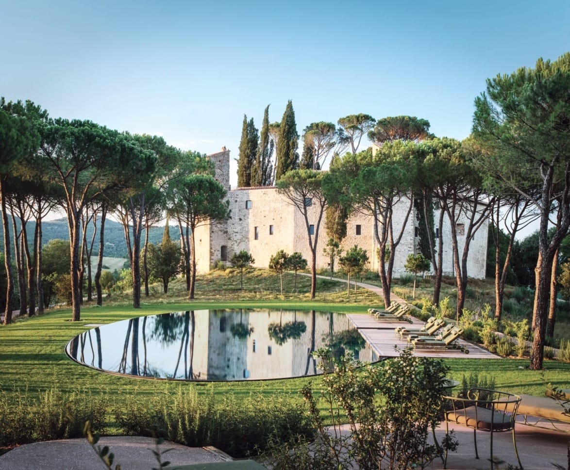 Scenic view of a historic stone castle surrounded by lush trees and circular pool in a serene landscape.
