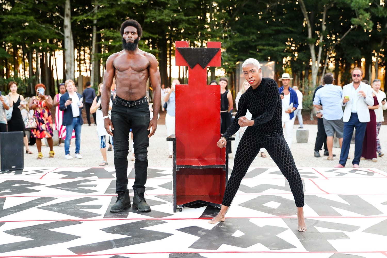 Two performers in a dynamic outdoor performance, one shirtless and the other in a black outfit, with a crowd in the background.