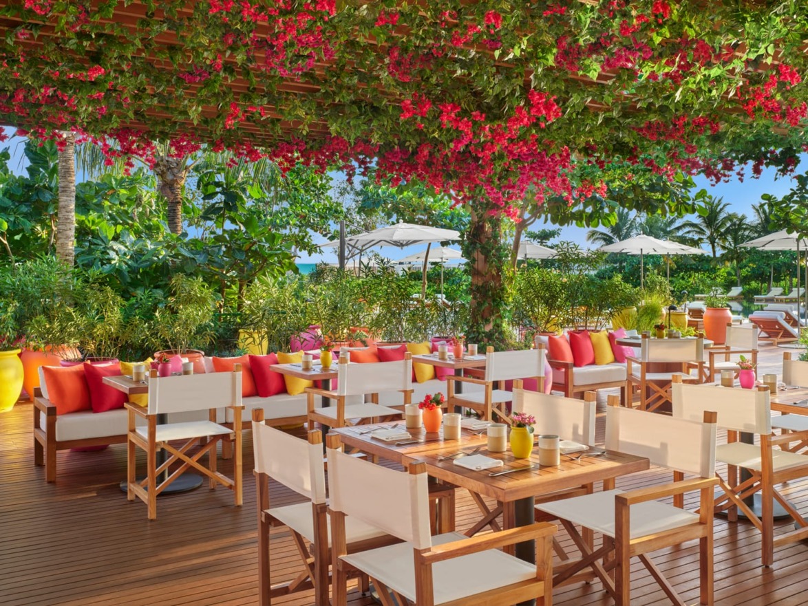 Outdoor restaurant patio with wooden tables, chairs, red cushions, and lush greenery under a canopy of vibrant flowers.