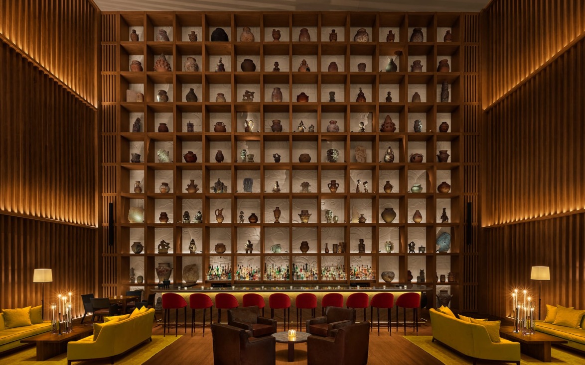 Cozy bar interior with warm lighting, large bookshelf filled with decorative pottery, yellow couches, and red barstools