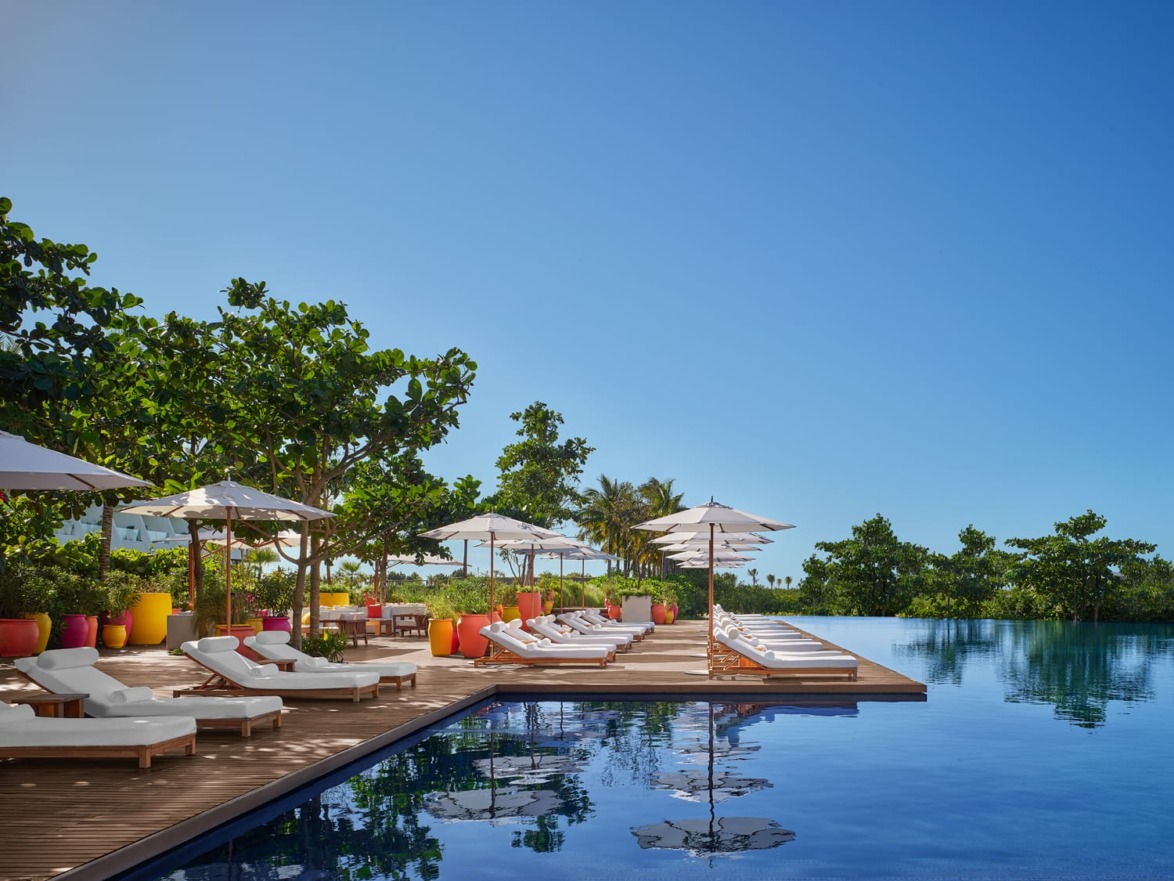 Hotel poolside area with sun loungers, umbrellas, and lush greenery under a clear blue sky.