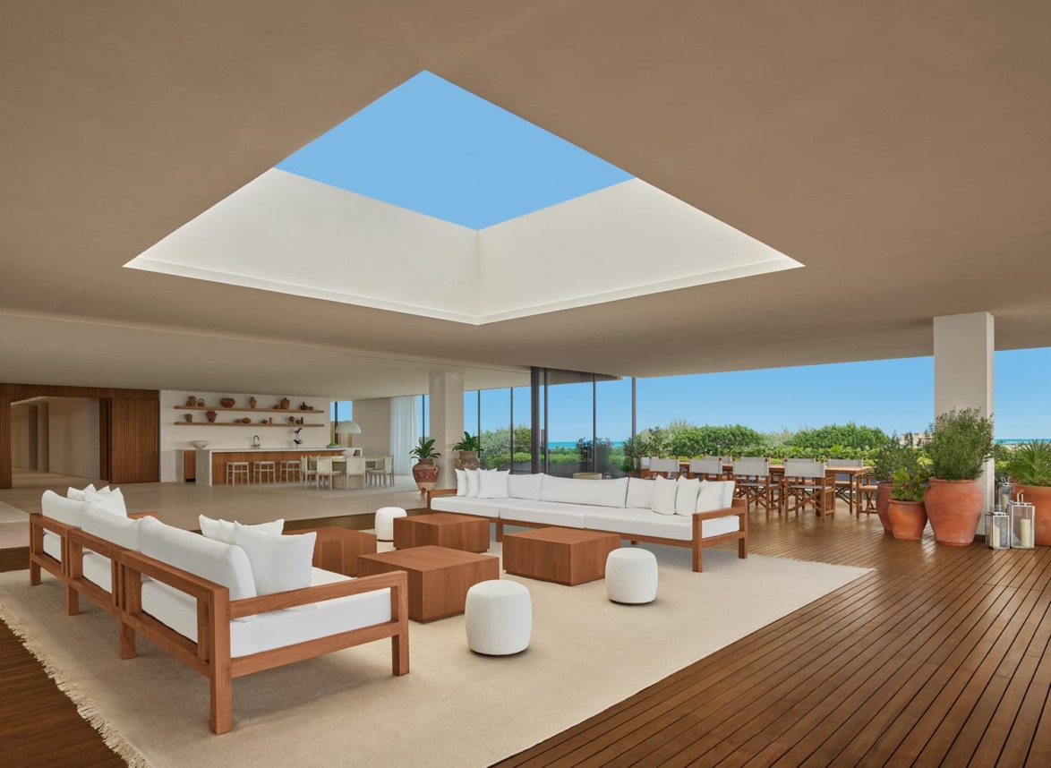 Spacious open terrace with modern wooden furniture, white sofas, a skylight, and views of lush greenery and a clear blue sky.
