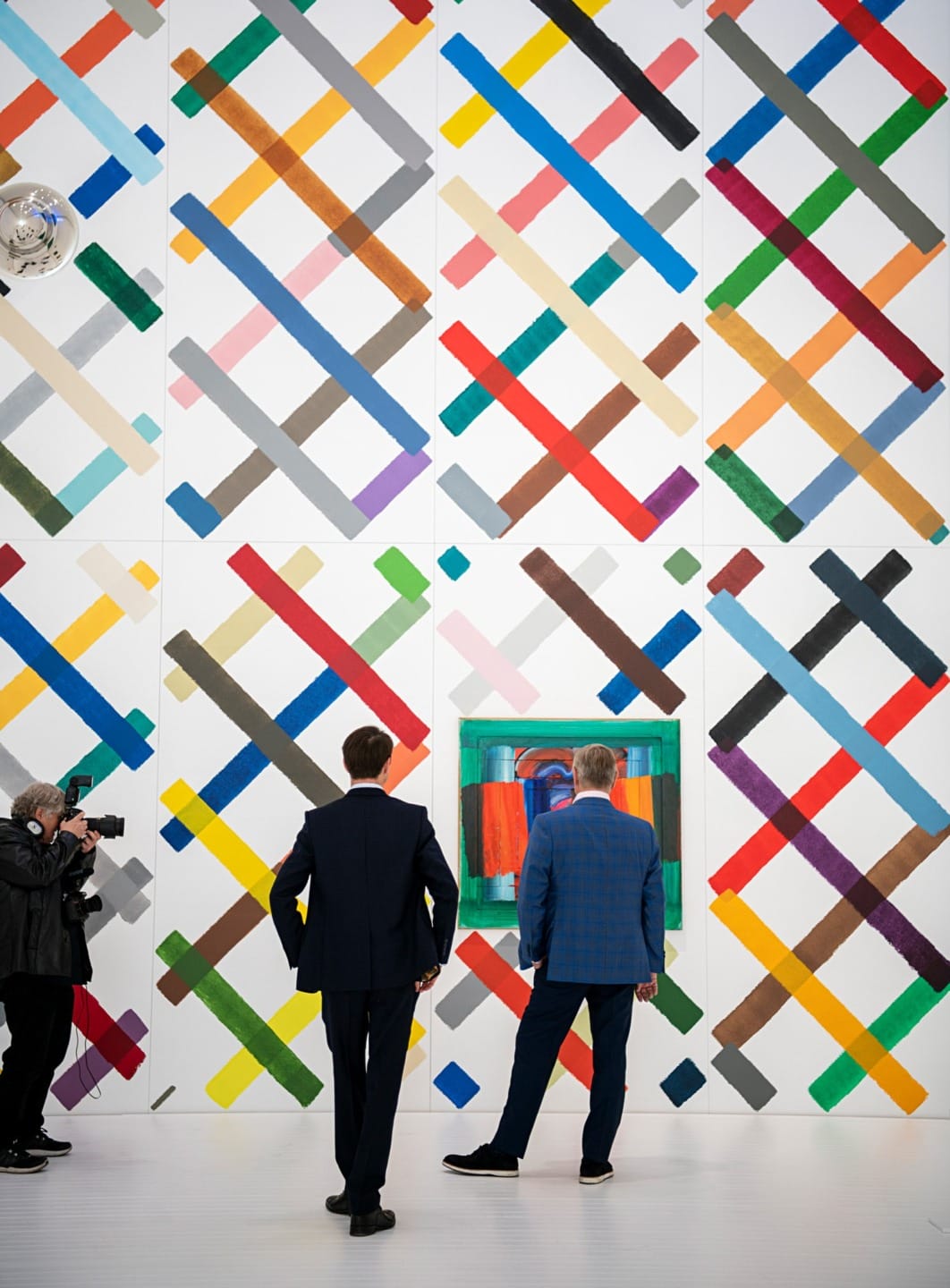 Two men stand in front of colorful geometric wall art while a photographer captures the scene in a bright gallery.