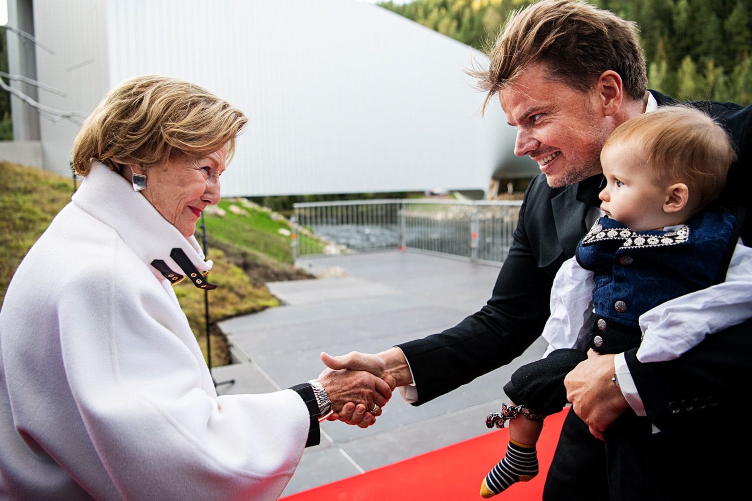 Woman in white coat shakes hands with a smiling man holding a baby on a red carpet event outdoors.
