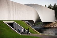 Modern architectural bridge with a unique twisting design over a river, surrounded by lush greenery and walking visitors.