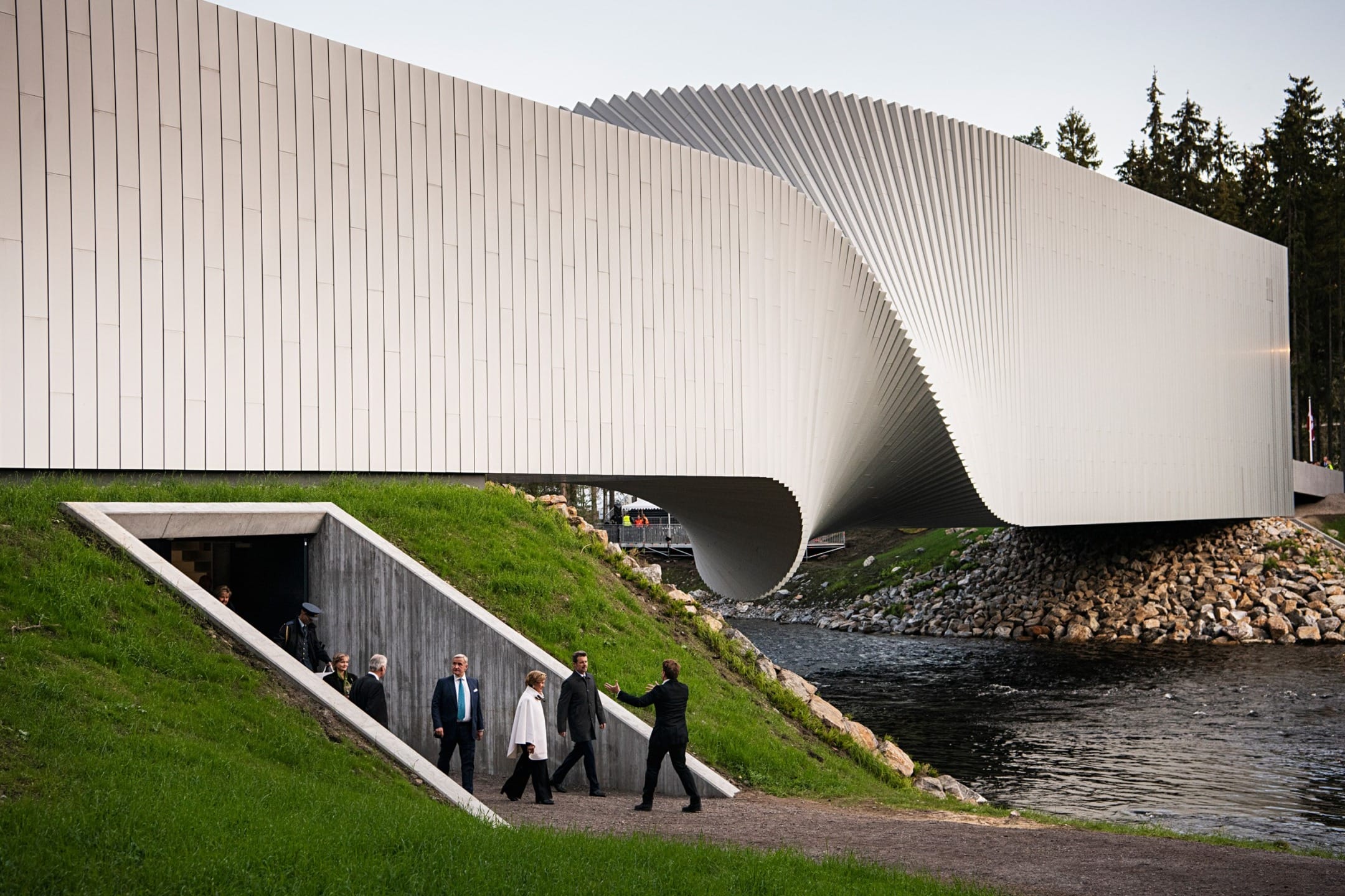 Modern architectural bridge with a unique twisting design over a river, surrounded by lush greenery and walking visitors.
