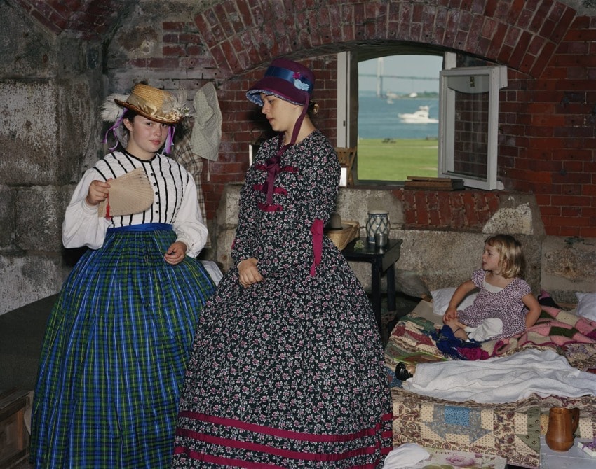 Women in historical dresses and hats in a stone room with a view of the sea, reading a paper near a child on a bed.