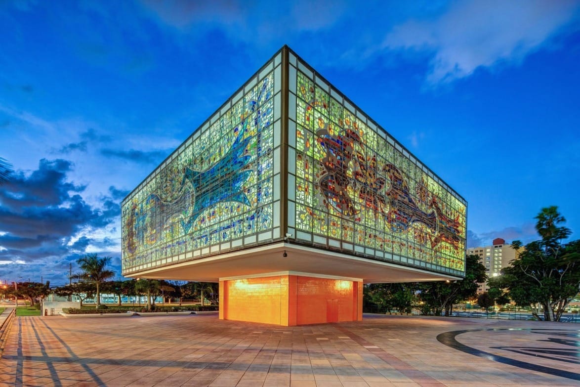 Glass-walled building with colorful stained glass designs, illuminated at twilight, set against a vibrant blue sky.