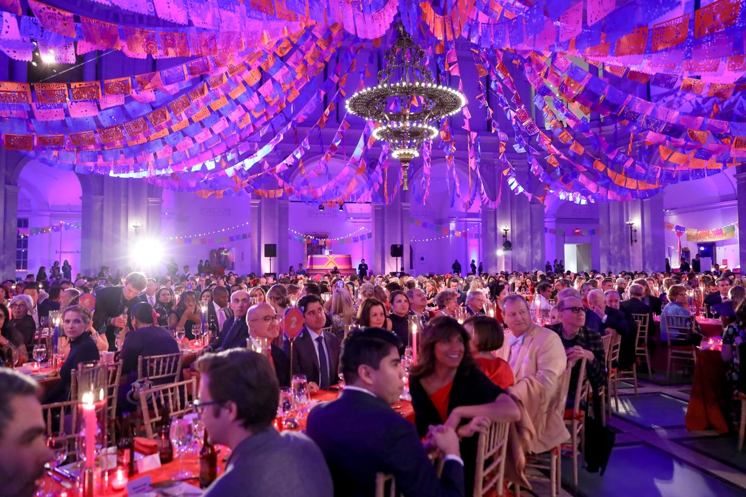 Elegant event in a grand hall with colorful decorations, a large crowd seated at tables under vibrant banners and chandeliers.