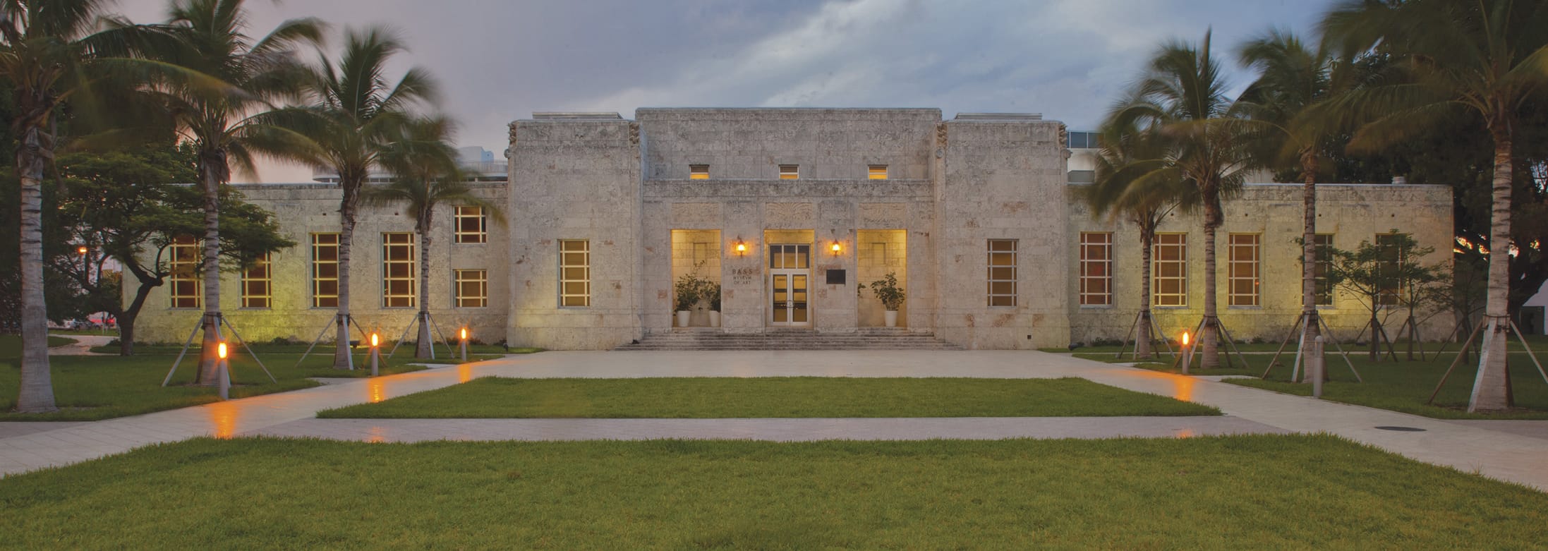 Art Deco building with palm trees lining a pathway leading to its illuminated entrance at dusk