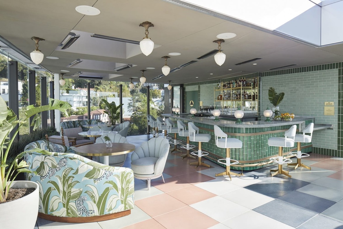 Rooftop bar with pastel decor, tropical print chairs, a green-tiled bar, and city views through large windows.
