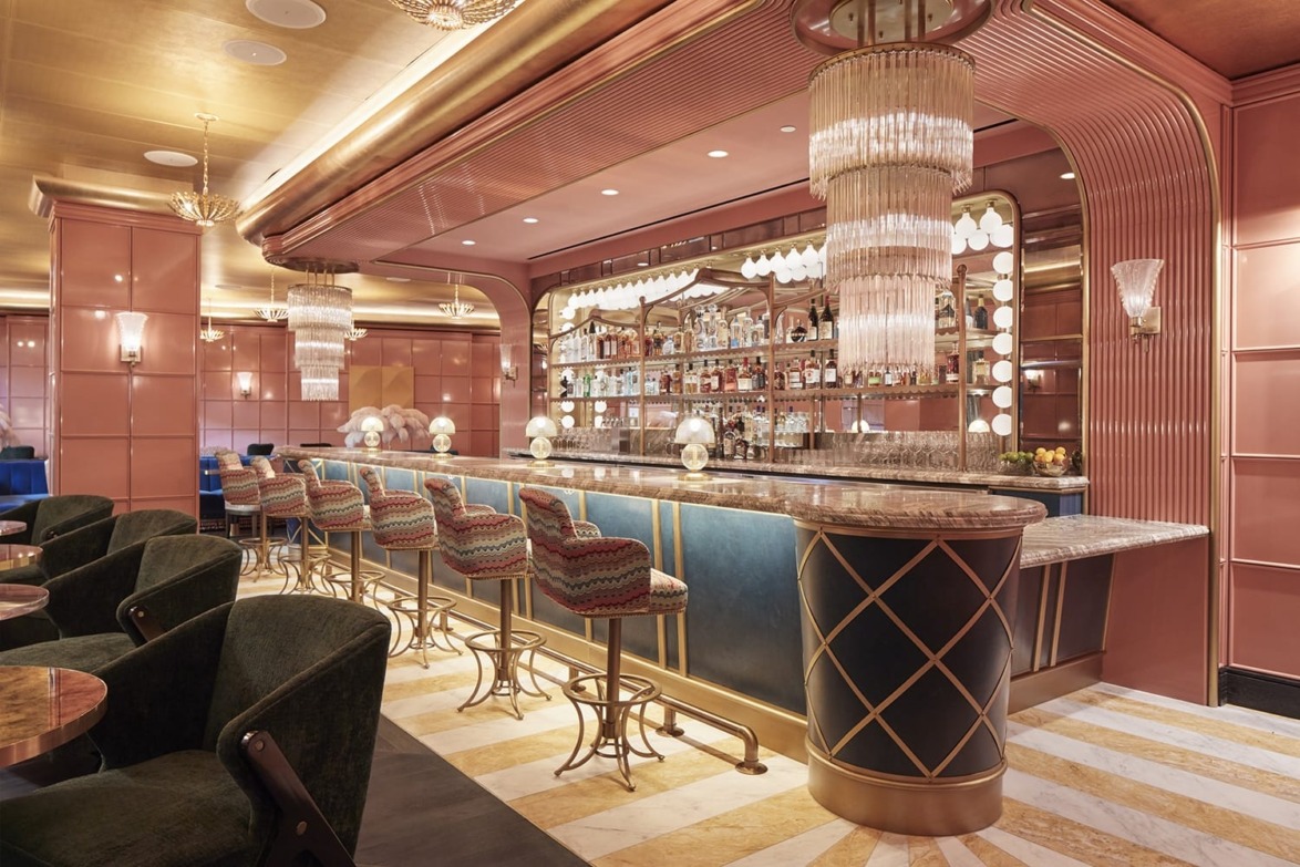 Art Deco-style bar interior with pink walls, elegant lighting, plush seating, and a marble countertop.