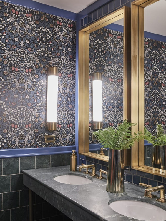 Bathroom with floral wallpaper, double sinks, gold-framed mirrors, elegant lighting fixtures, and a vase with green ferns.