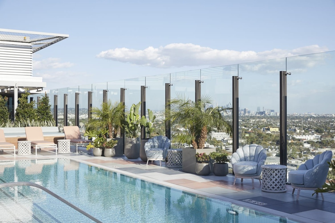 Rooftop pool area with glass railing, plants, and seating, overlooking city skyline under a blue sky with scattered clouds.