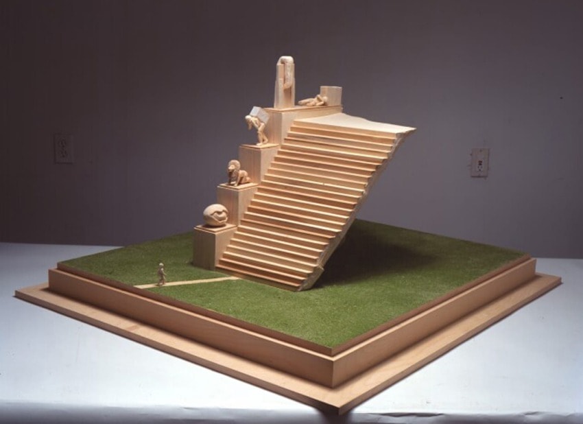 Miniature sculpture of grand staircase with human figures on artificial grass platform.