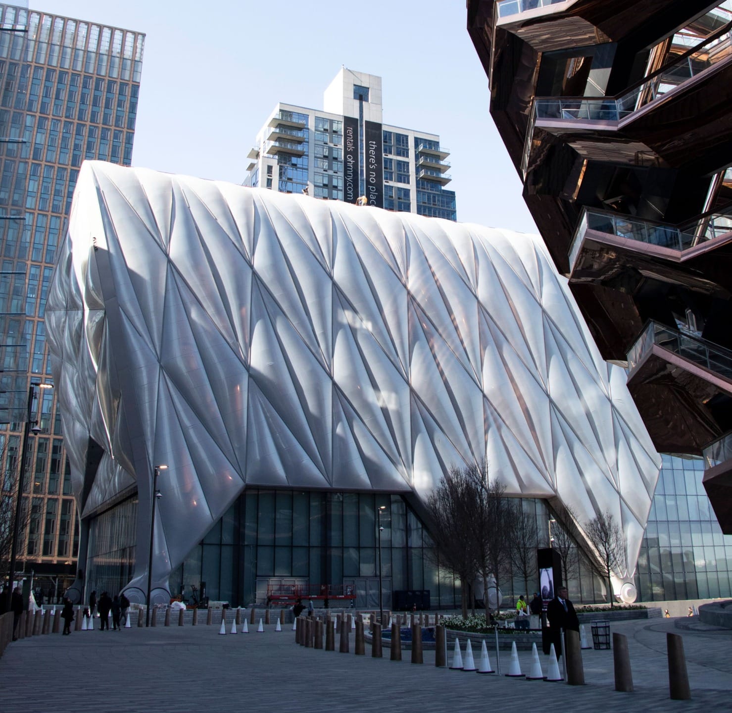 Modern architectural building with a geometric facade in an urban setting surrounded by skyscrapers and outdoor walkway.
