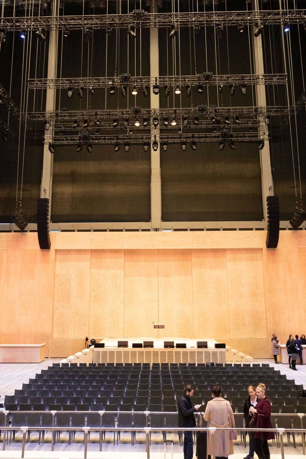 Empty auditorium with rows of black chairs, large wooden stage, and overhead lighting; a few people stand in conversation.