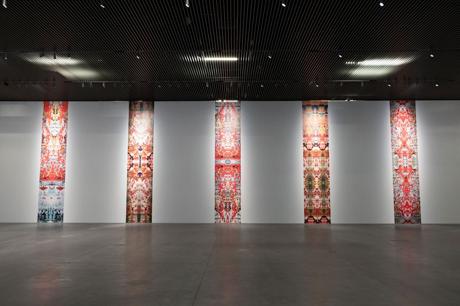 Six vibrant, vertically hung tapestries with intricate patterns displayed in a modern gallery setting on a gray concrete floor.