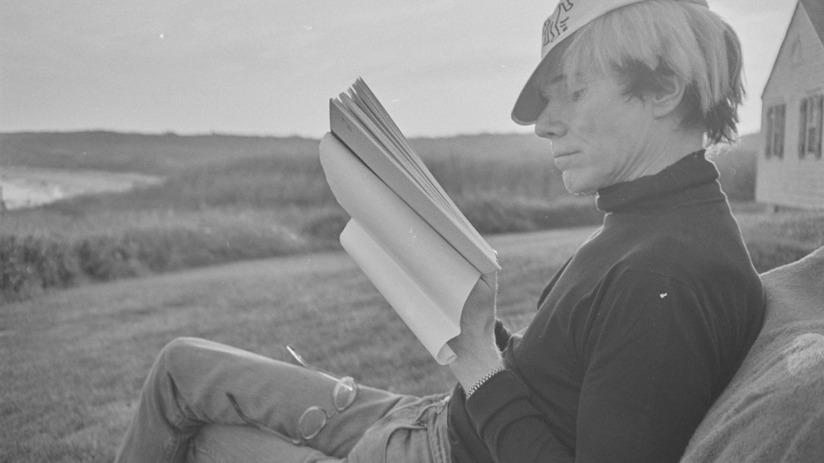 Person sitting outdoors reading a book with a hat, glasses on lap, nature background.