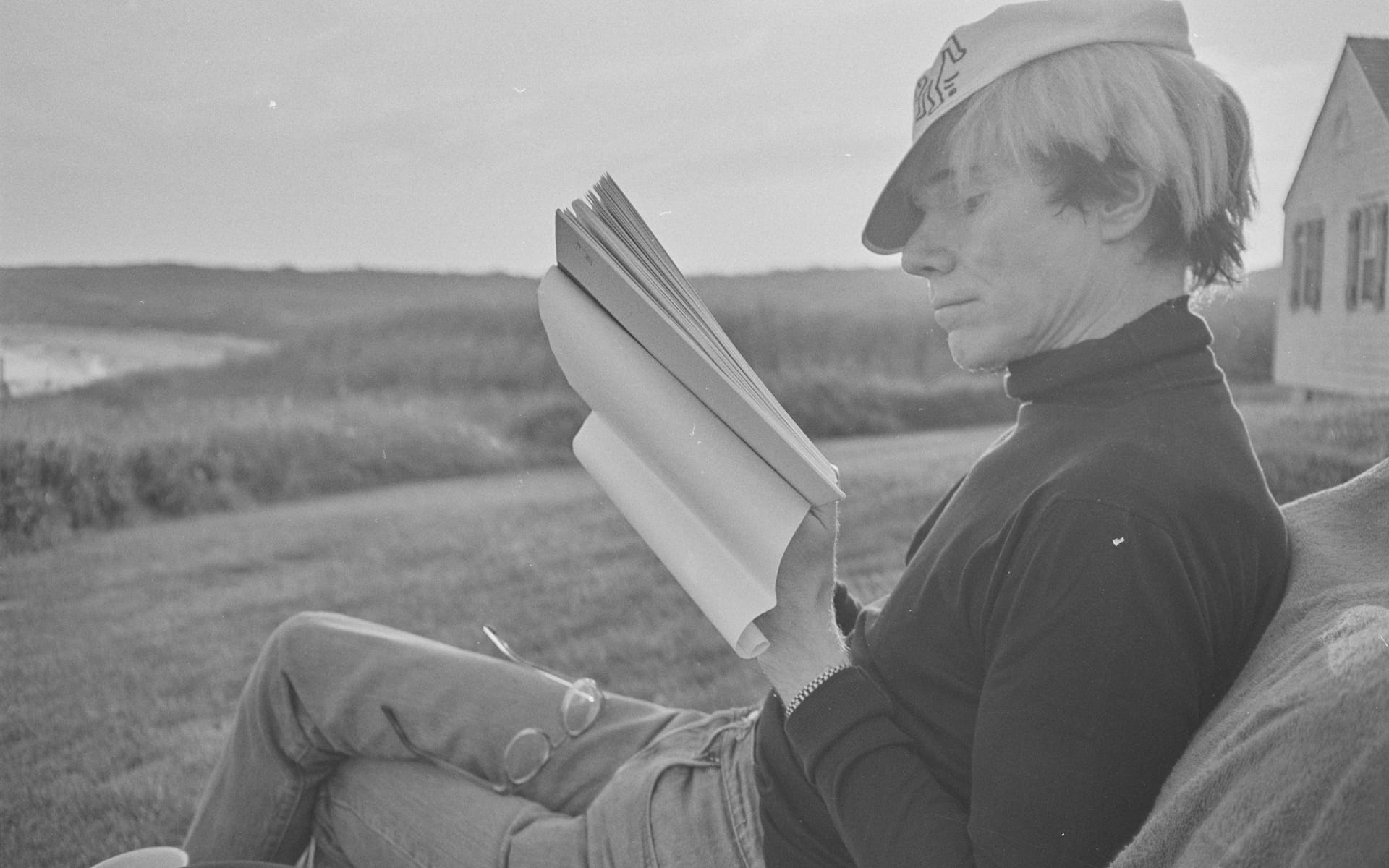 Person sitting outdoors reading a book with a hat, glasses on lap, nature background.