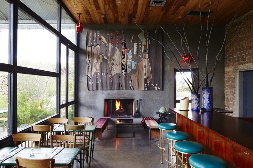 Cozy cafe interior with a fireplace, colorful bar stools, and a tapestry on the wall, surrounded by large windows.