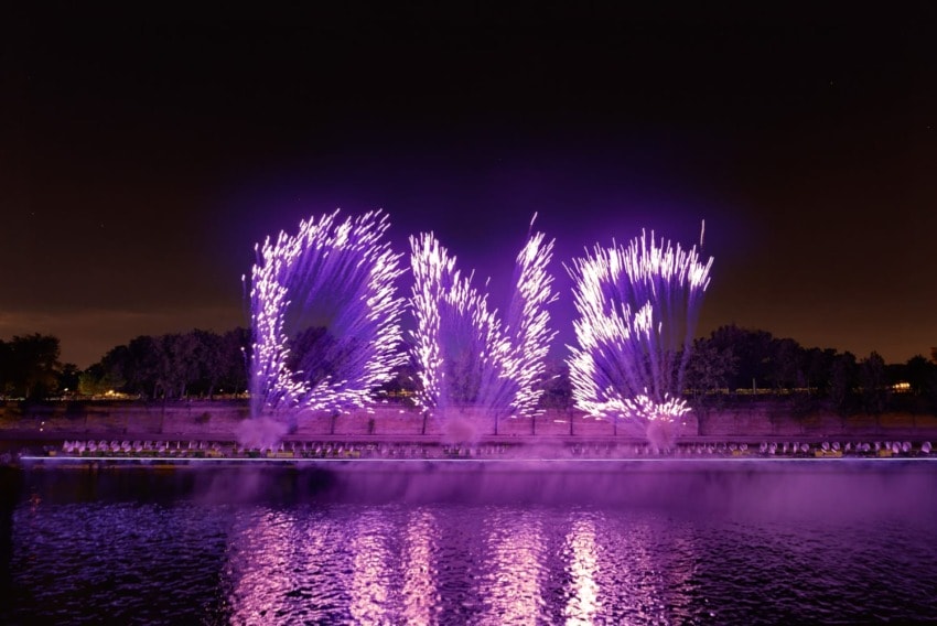 Purple fireworks spelling "LOVE" reflect on a body of water at night with trees in the background.