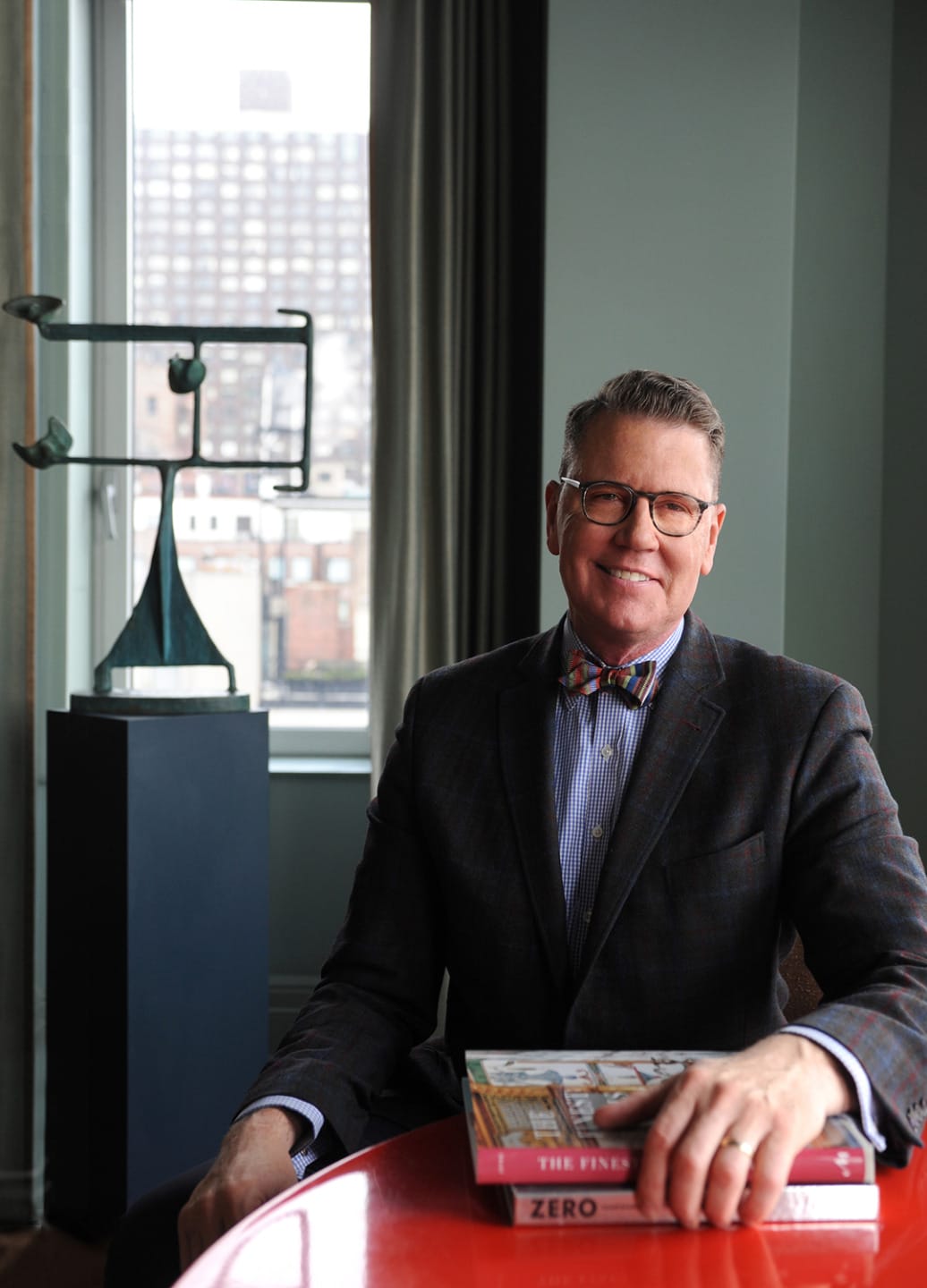 Man sitting at a red table with books, smiling, wearing glasses and a suit, with a sculpture and city view in the background.