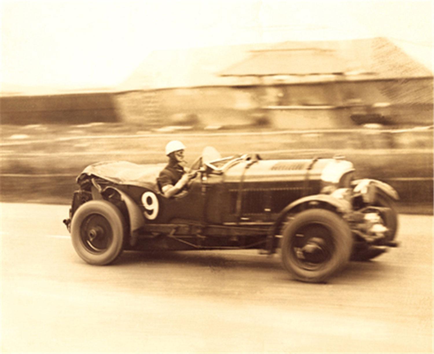 Vintage race car with driver wearing a helmet, speeding on a road, blurred background of buildings and landscape.
