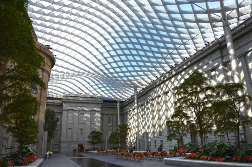Glass-ceilinged atrium with trees and cafe seating, inside a classic stone building.