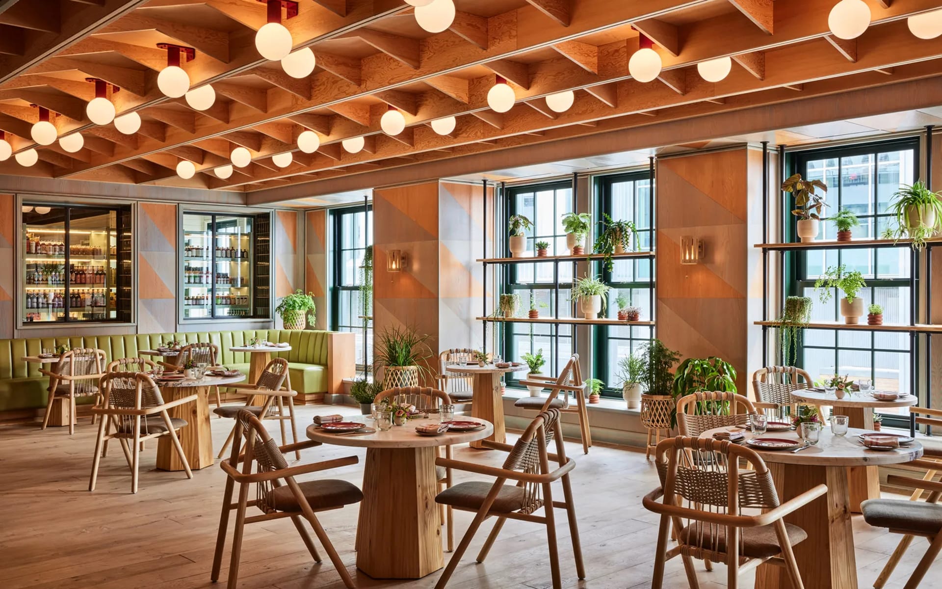 Modern restaurant interior with wooden furniture, large windows, and hanging lights. Tables are set and potted plants decorate shelves.