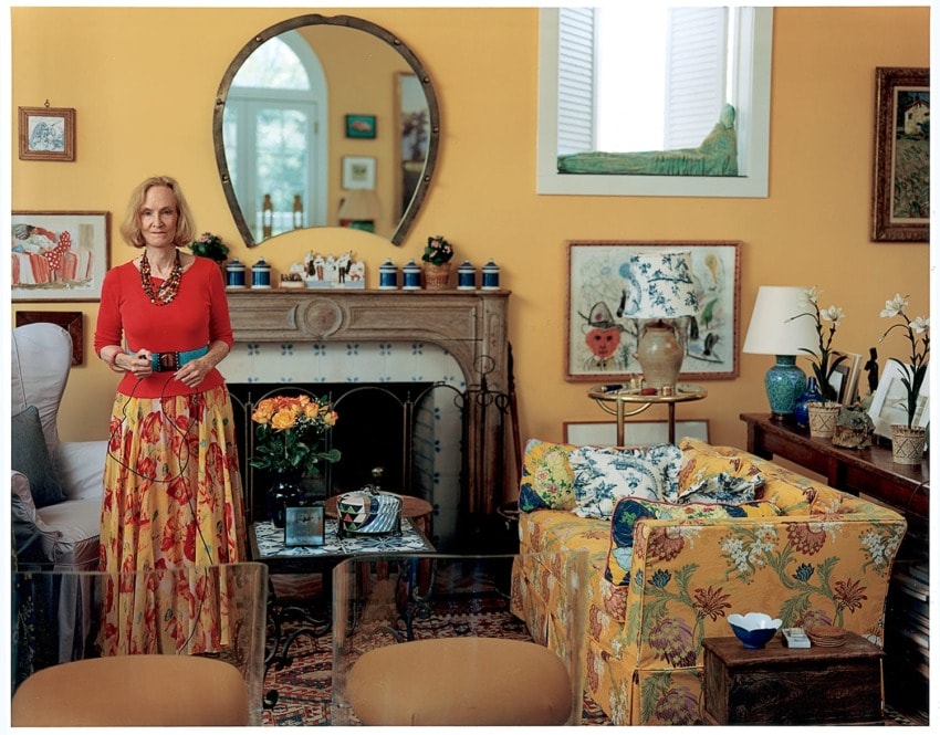 A woman stands in a colorful, art-filled living room with a round mirror and floral patterns throughout.