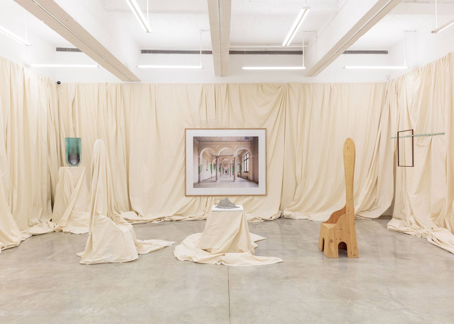 Contemporary art installation with draped fabric, framed artwork, wooden chair, and ceramic sculptures in a gallery setting.