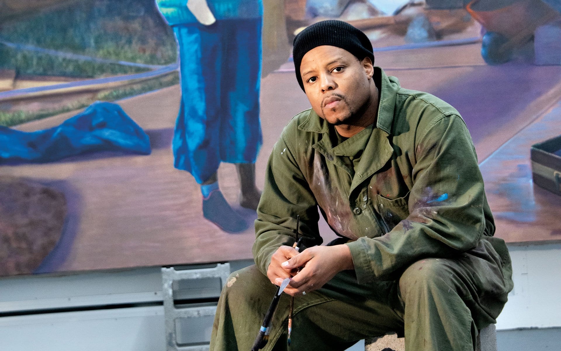 Artist in a green jumpsuit sitting with a paintbrush, colorful mural in the background.
